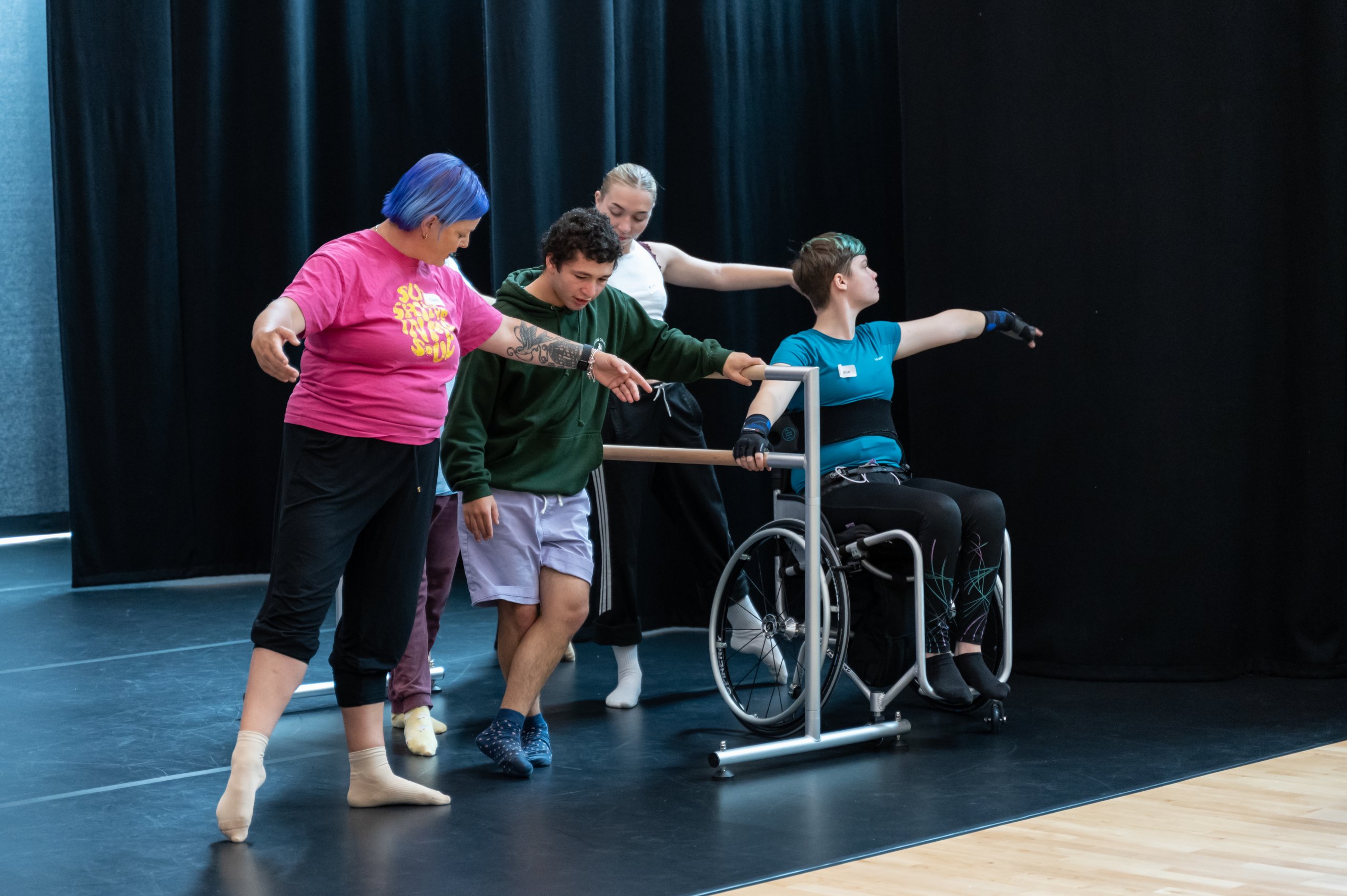 Four dancers around a ballet barre with arms extended out to the side. One dancer in a pink t-shirt is not touching the barre, a dancer in a wheelchair and two other dancers are holding the barre with one arm. 