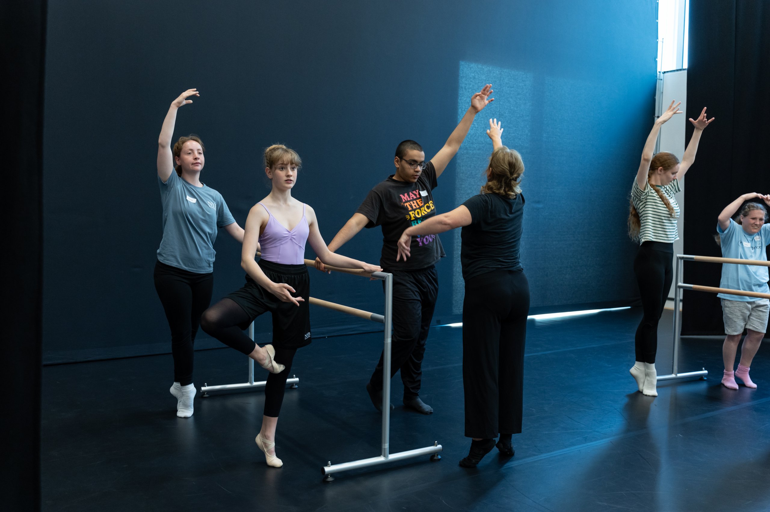 Six dancers surround a ballet barre, choosing different ways to reach and balance. 