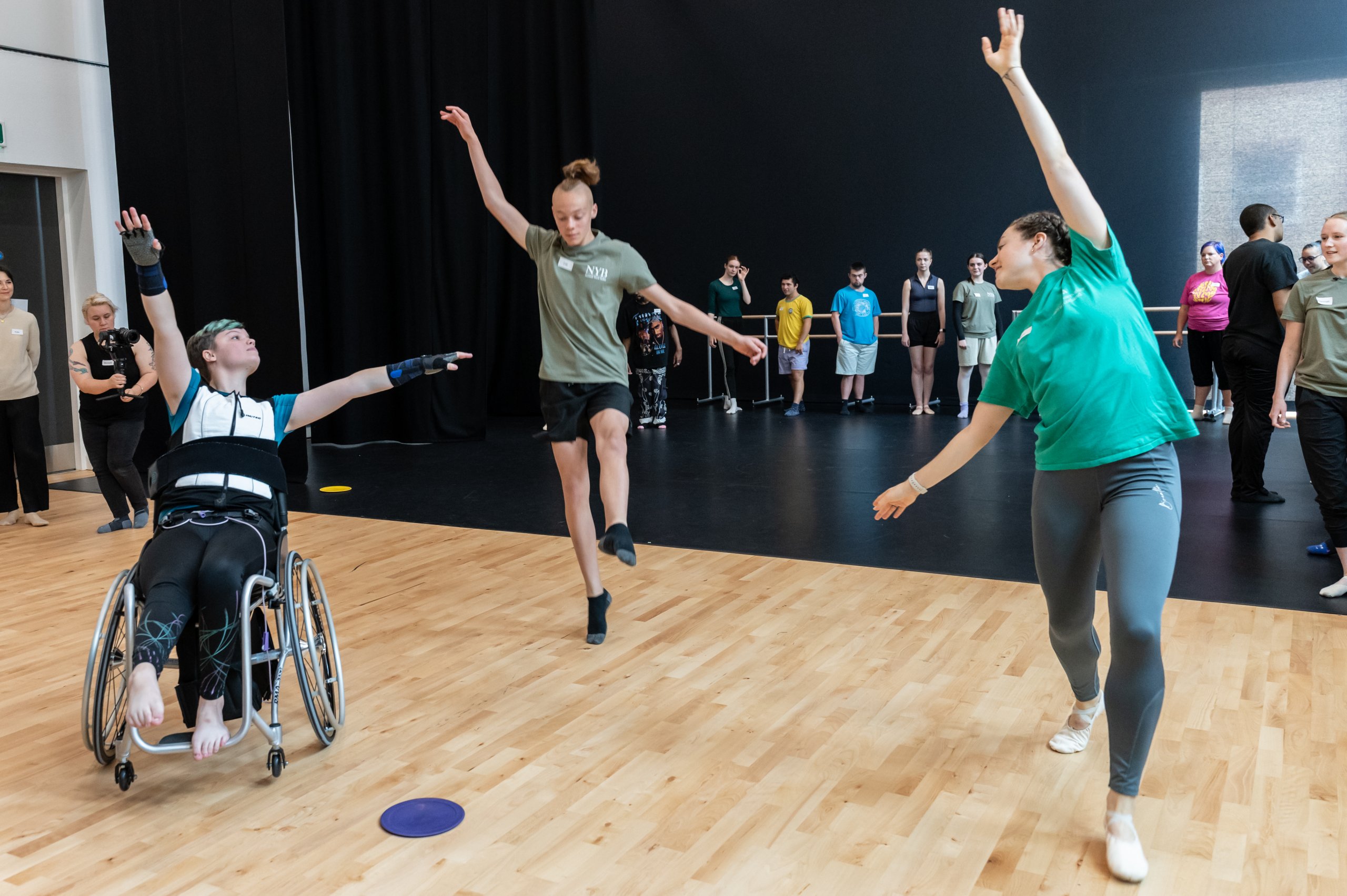 A dancer in a wheelchair gliding forward with one arm reaching upwards and the other out to the side by two dancers with arms reaching beside them diagonally. Several dancers are observing in the background. 