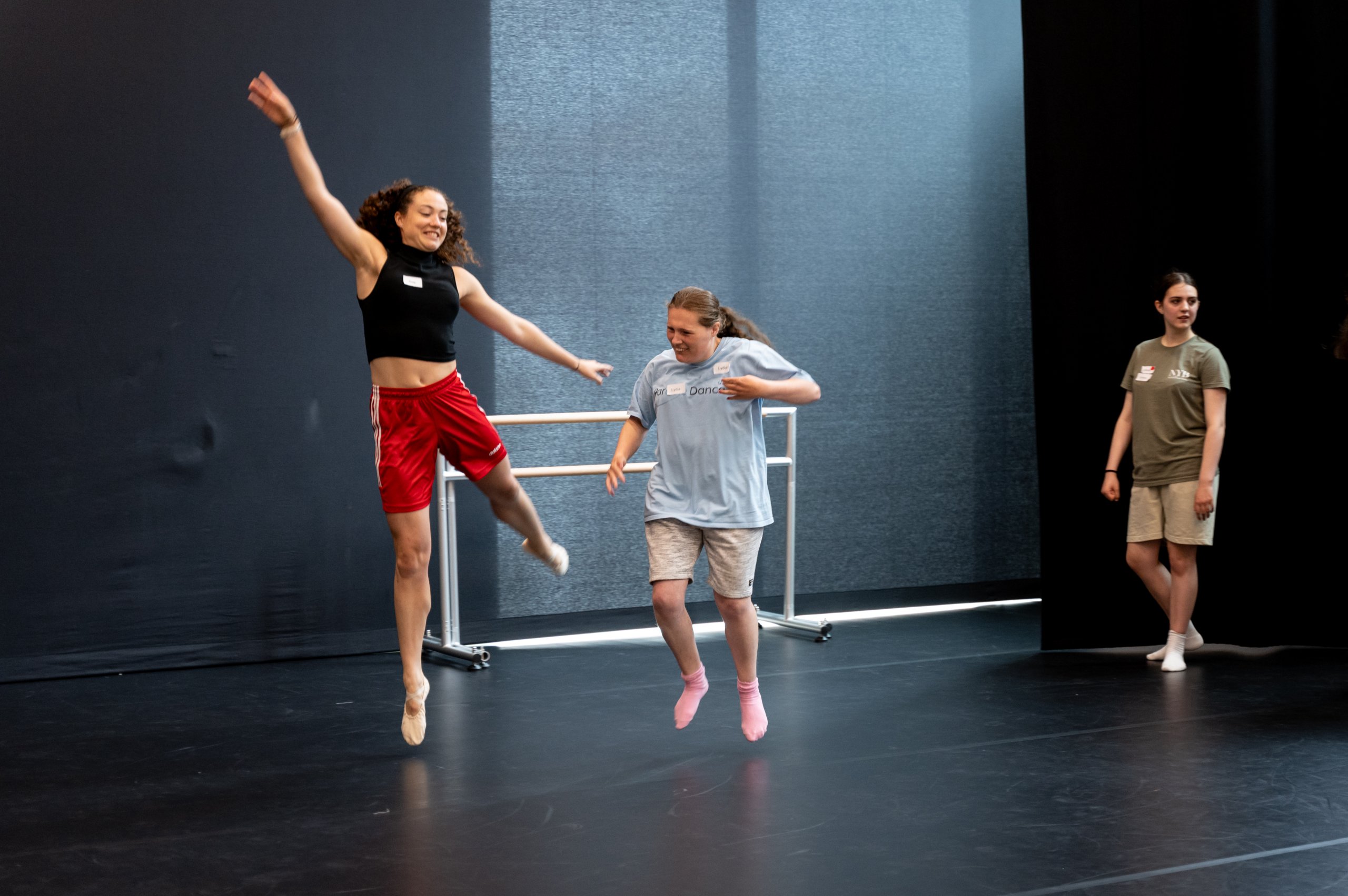 Two dancers are elevating side by side. One dancer has their leg extended behind them and another is jumping with their legs slightly apart. There is a dancer and a ballet barre in the background. 