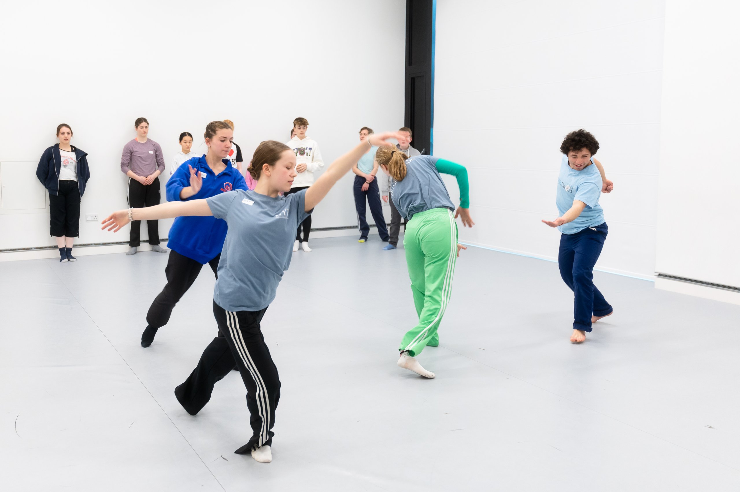 Many dancers sharing the studio space in a running motion, travelling in different directions with some dancers observing in the background. 