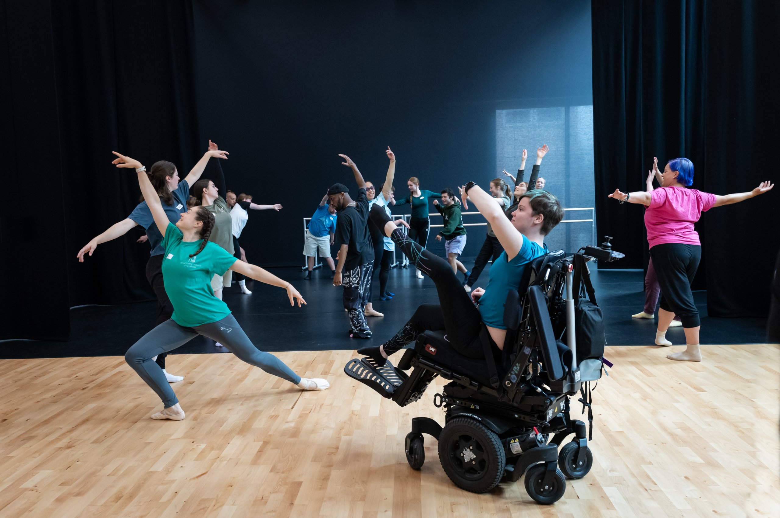 Whole company of dancers finding length in their bodies. The studio is filled with dancers who are standing, some are lunging, a dancer in a wheelchair is seated. 