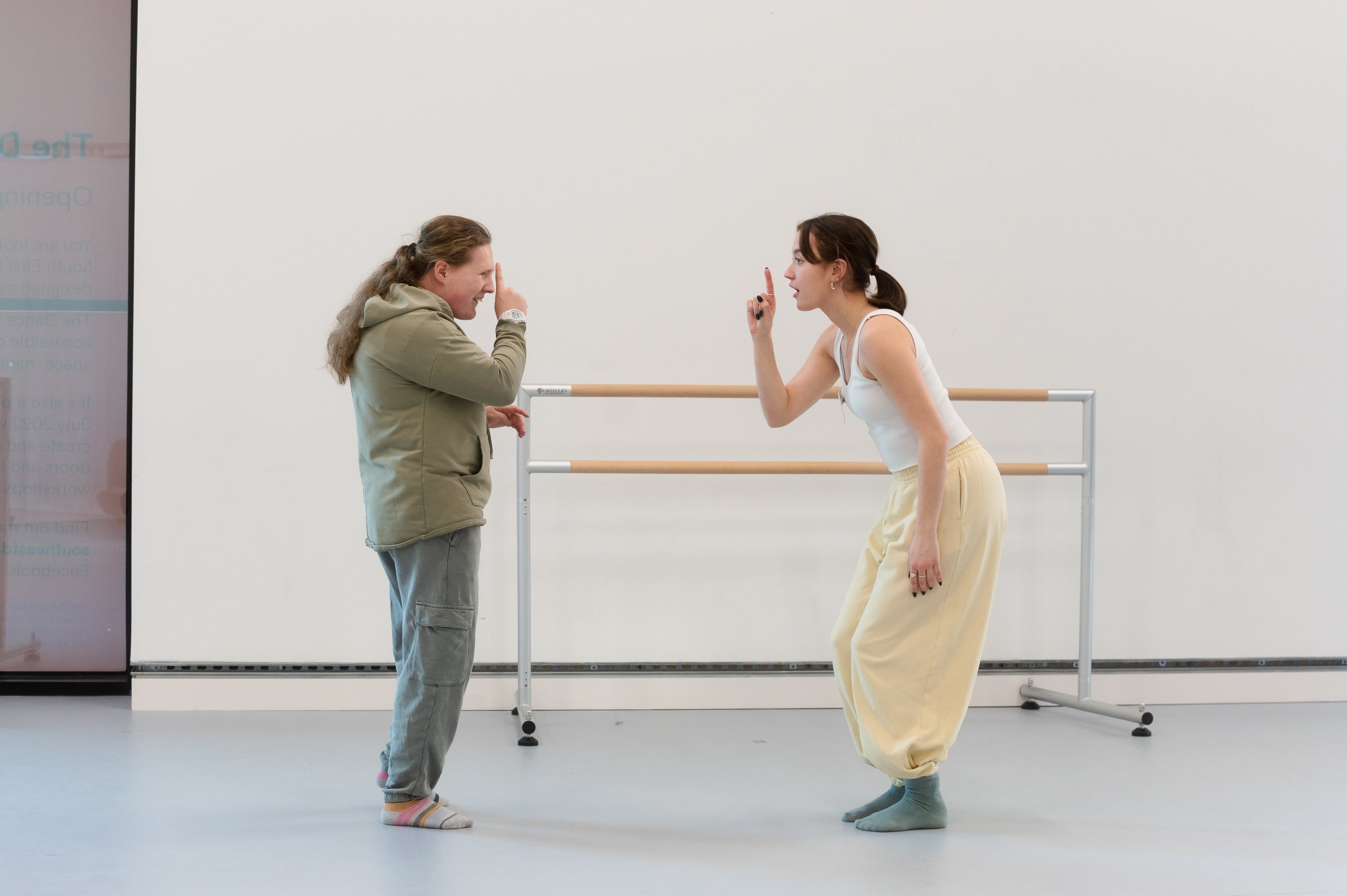 Two dancers facing each other with their feet in parallel with a finger pointing upwards directly in front of their faces. There is a ballet barre in the background. 