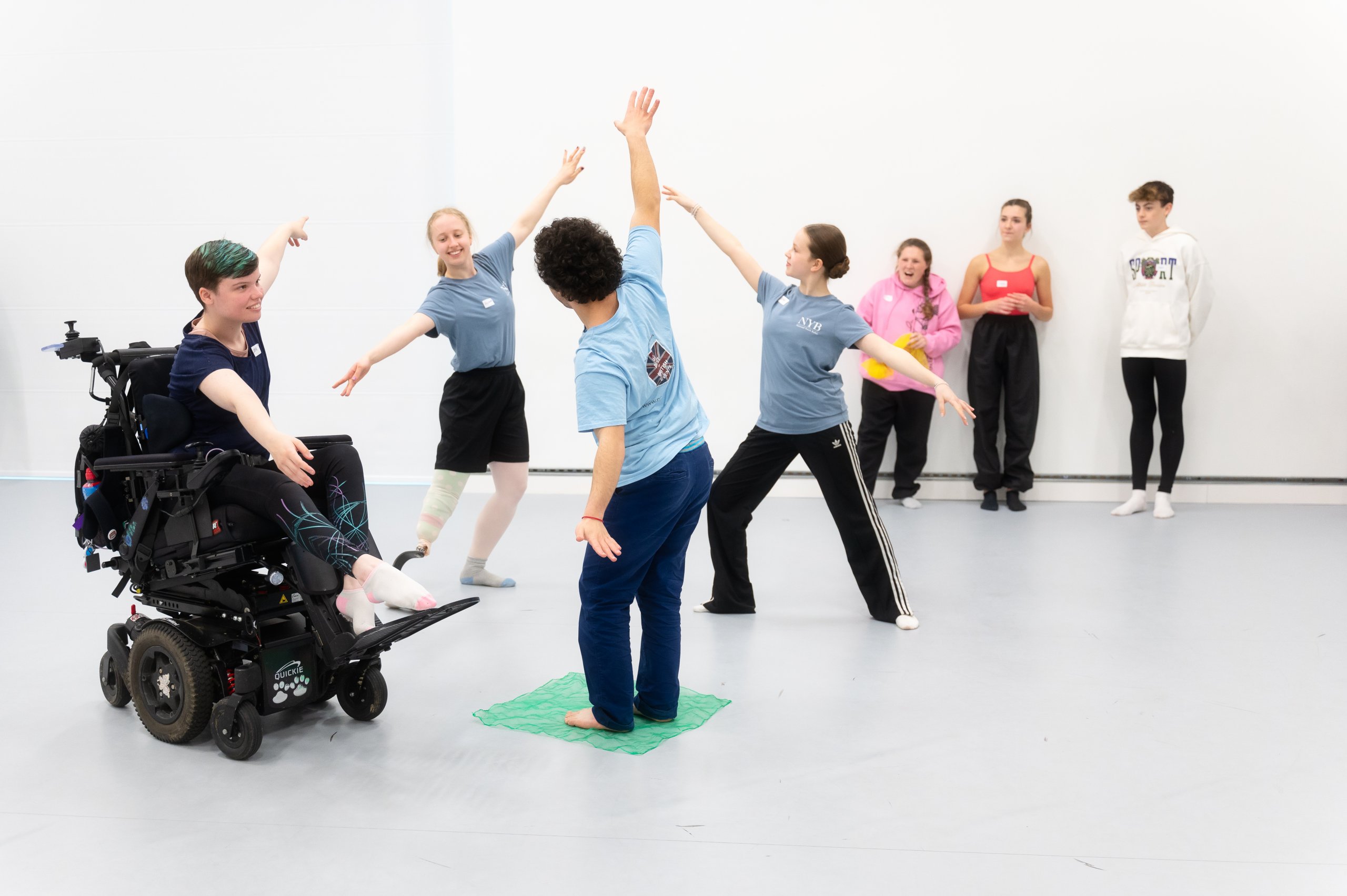 One dancer stood on a green piece of material, another is sat in a wheelchair, and two more are lunging. These dancers have their arms out on a diagonal whilst other dancers observe in the background. 