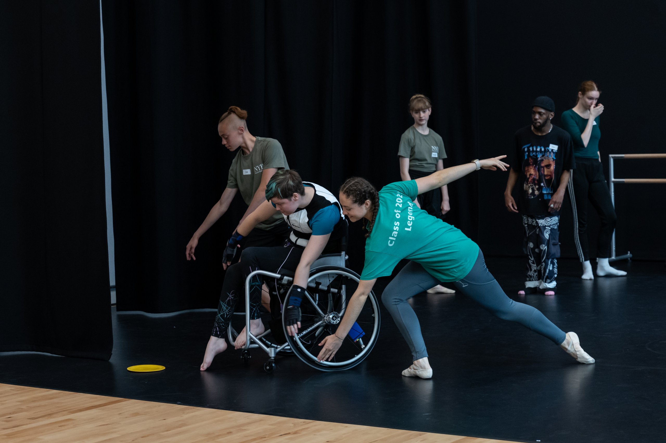 One dancer reaching arms diagonally low next to a dancer in a wheelchair reaching forwards and low next to another dancer lunging with arms extending diagonally away from each other. There are 3 people behind observing this. 