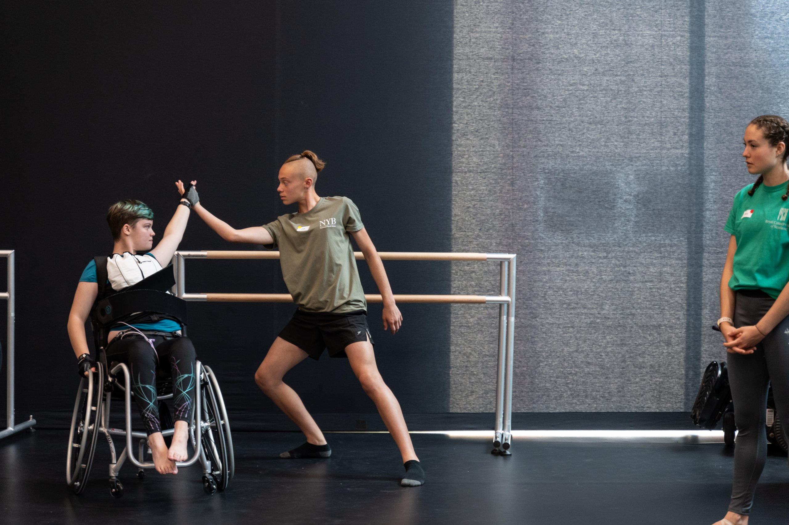 A dancer in a wheelchair touches hands with a dancer lunging whilst another person watches from the side. 