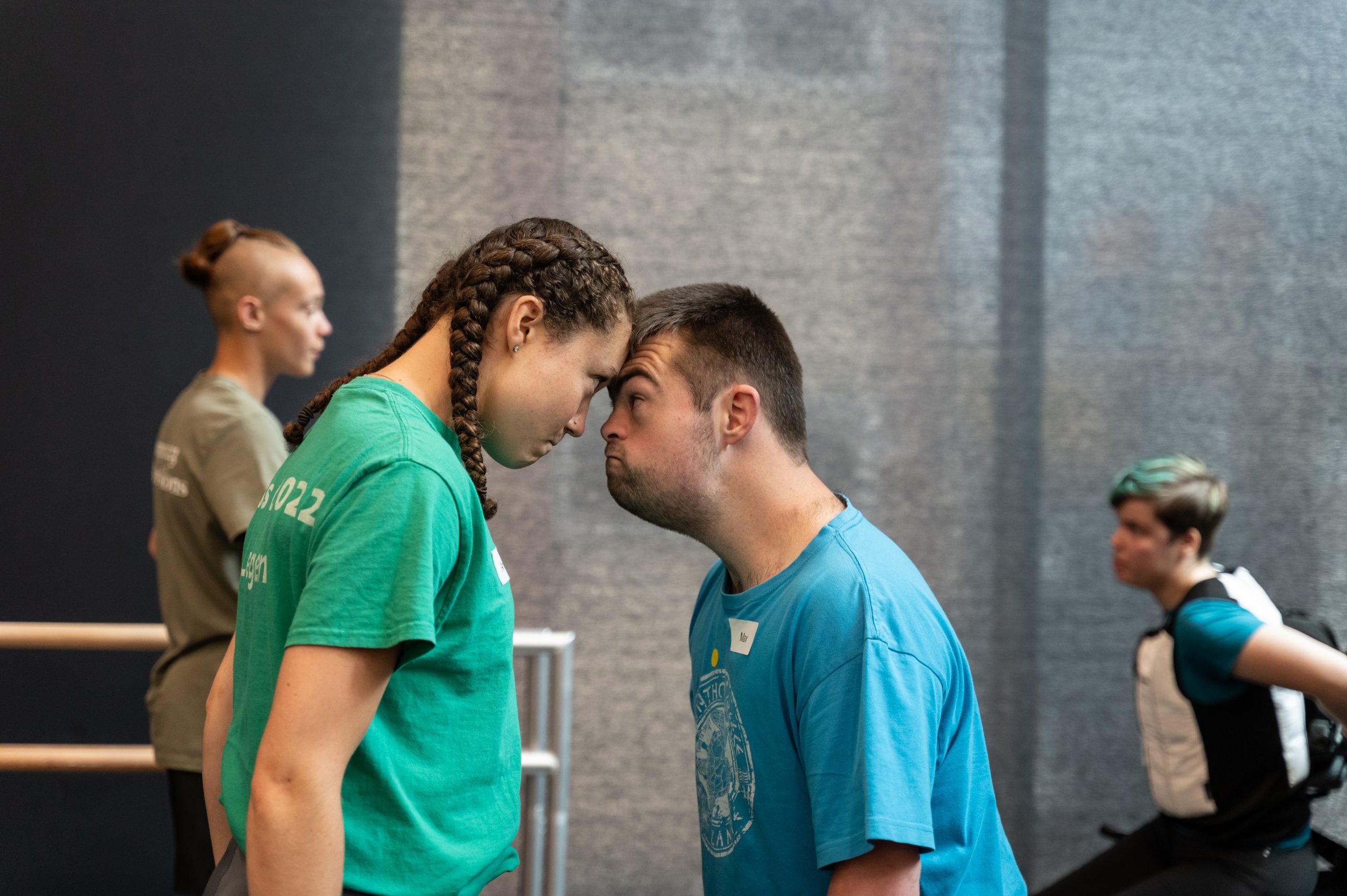 Two dancers making contact with their foreheads and making eye contact. One dancer is in a blue t-shirt and another in a green t-shirt. There are two people dancing in the background. 