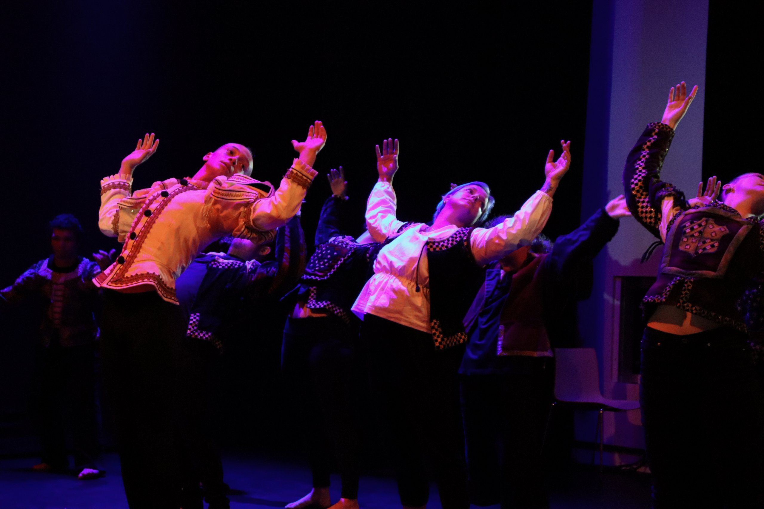 A group of dancers in costume, on stage leaning backwards with arms bent at the elbow and raised in line with their heads. 