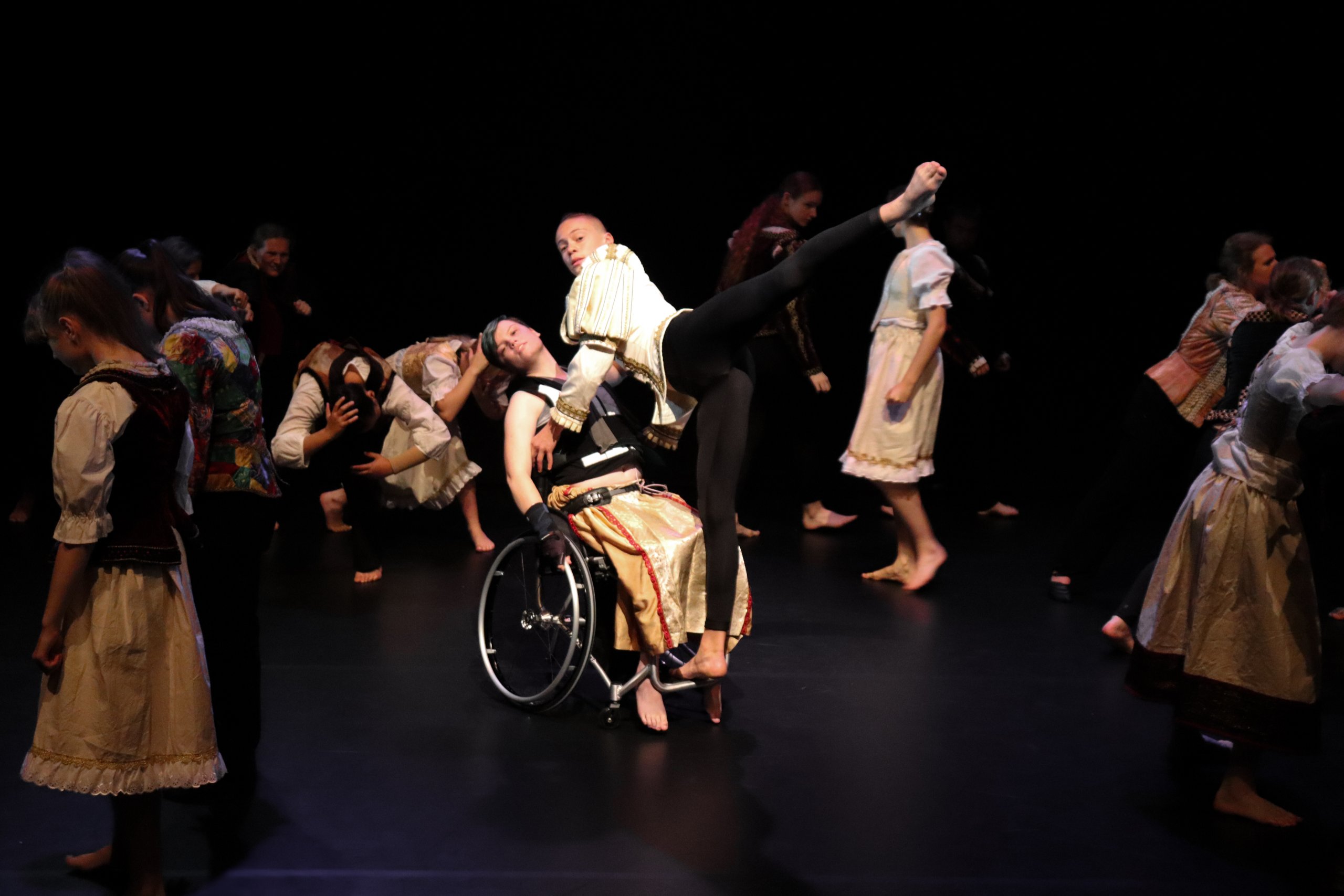 A dancer with their leg extended to the back leaning and balancing on a wheelchair whilst the dancer in the wheelchair leans backwards. Other dancers surround the space around them. 