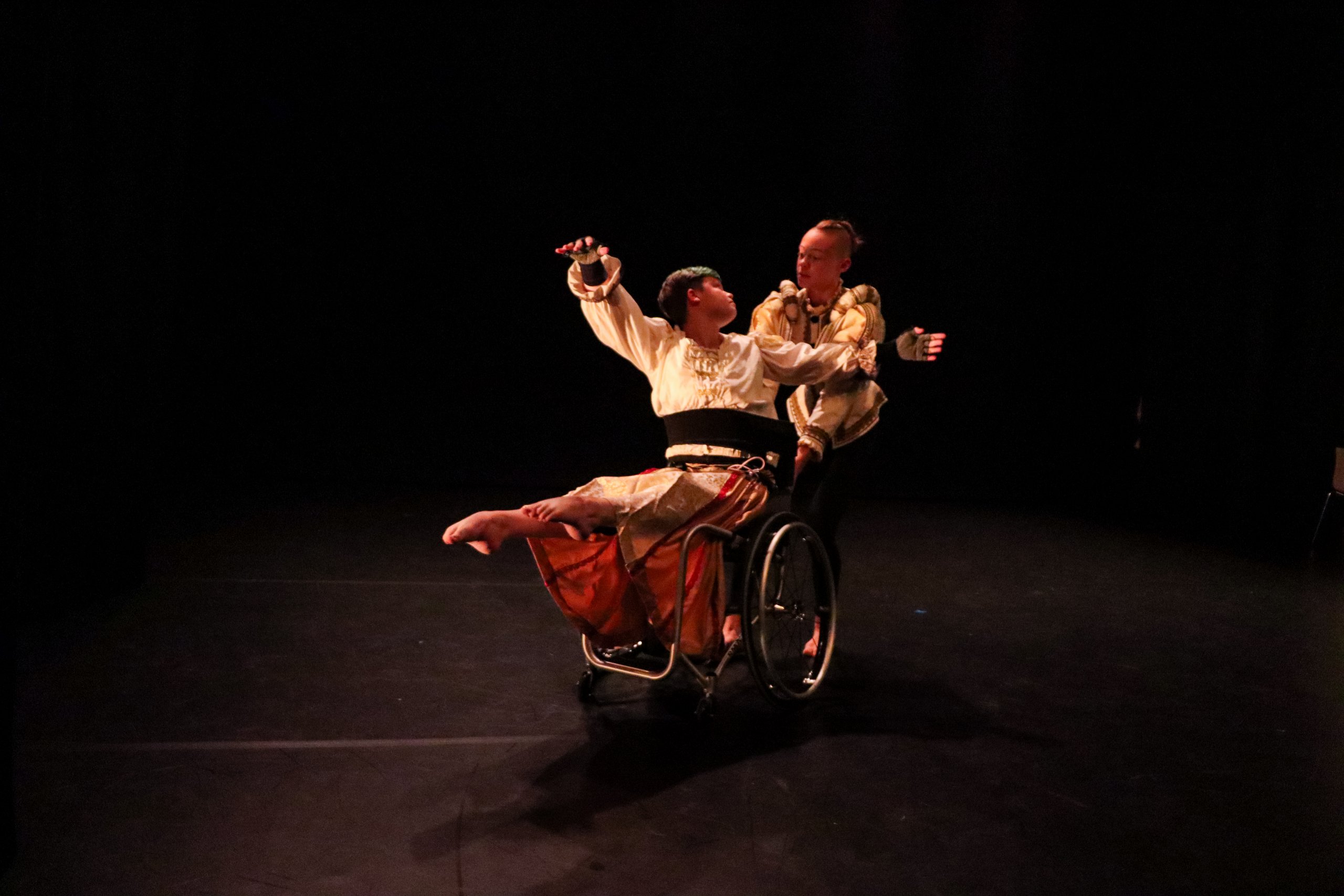 A dancer pulling another dancer in a wheelchair whilst they lean back with legs extended out. 