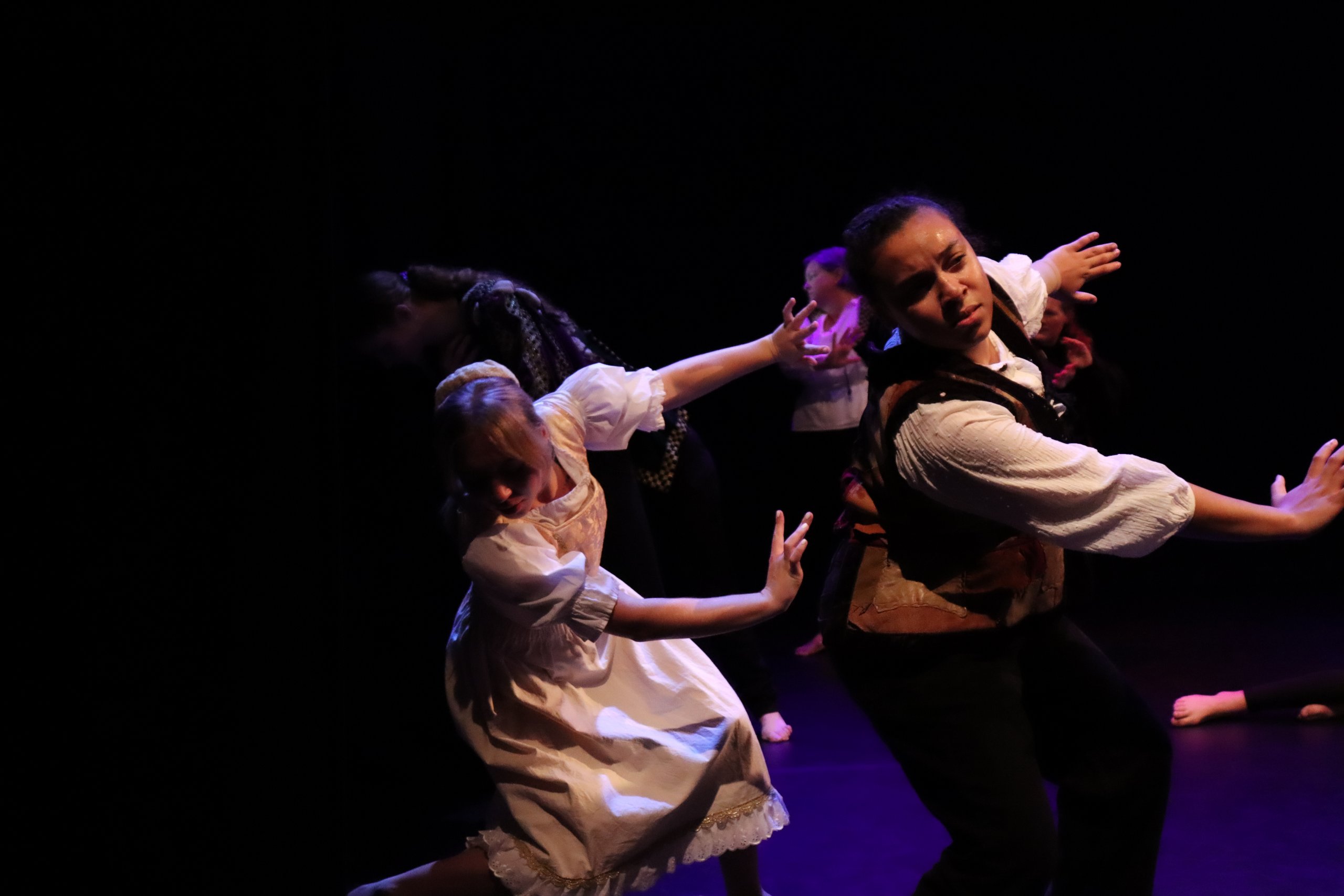 Two dancers in costume lit from the side with both arms extended to one side with their heads looking the opposite direction. 